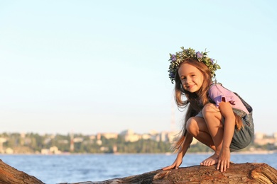 Cute little girl wearing flower wreath on tree near river, space for text. Child spending time in nature