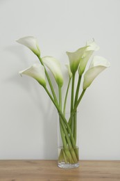 Photo of Beautiful calla lily flowers in glass vase on wooden table near white wall