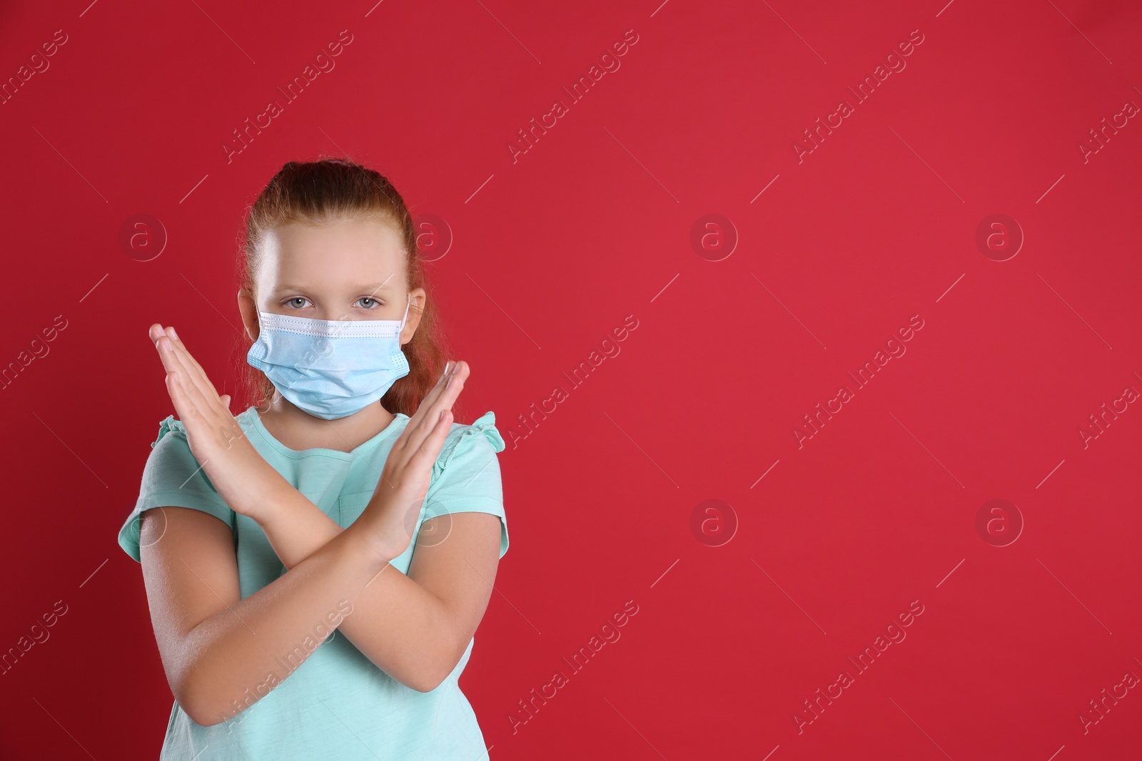 Photo of Little girl in protective mask showing stop gesture on red background, space for text. Prevent spreading of coronavirus