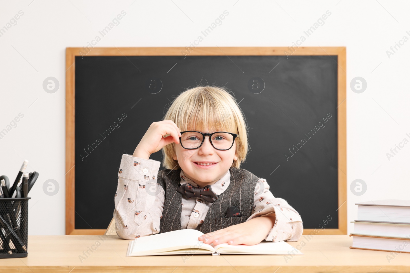 Photo of Happy little school child sitting at desk with books near chalkboard in classroom