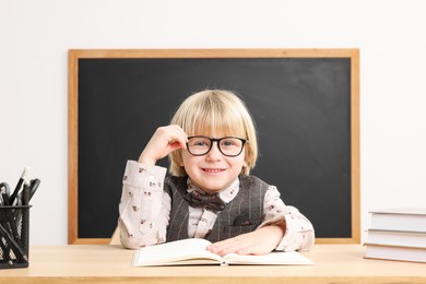 Happy little school child sitting at desk with books near chalkboard in classroom