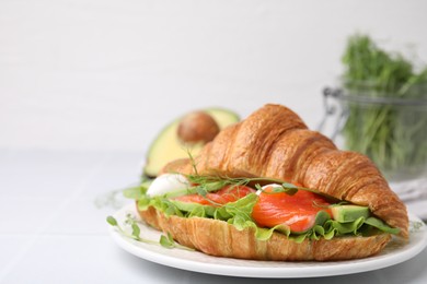 Tasty croissant with salmon, avocado, mozzarella and lettuce on white tiled table, closeup. Space for text