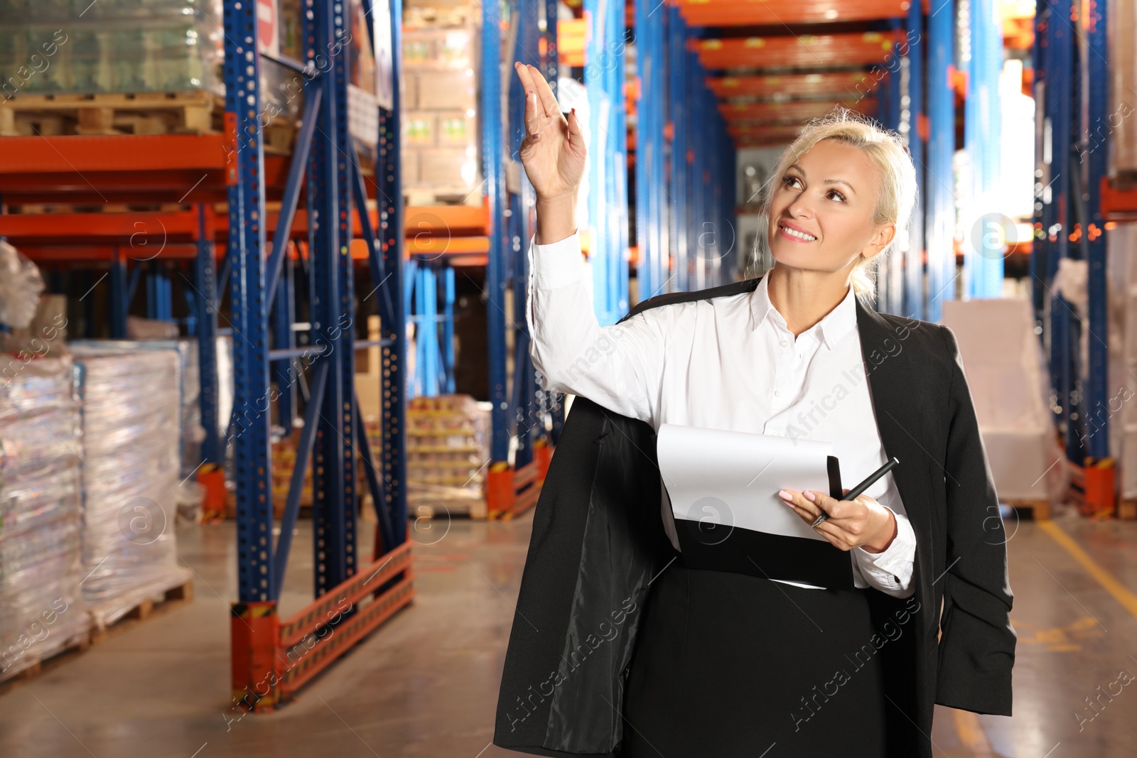 Photo of Happy manager holding clipboard and pointing at something in warehouse with lots of products