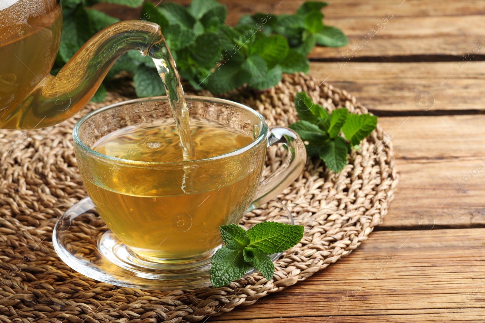 Photo of Pouring hot aromatic mint tea into cup at wooden table. Space for text