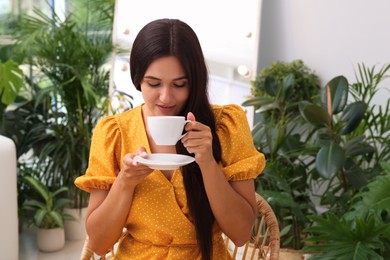 Beautiful woman with cup sitting in wicker armchair near houseplants indoors. Interior design