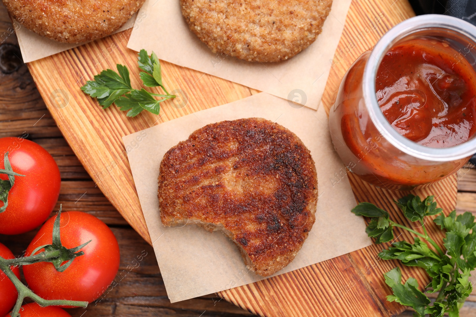 Photo of Delicious bitten vegan cutlets, ketchup, parsley and tomatoes on wooden table