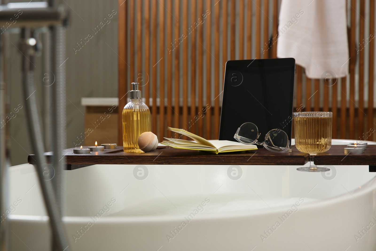 Photo of Wooden bath tray with candles, tablet, book and glass of drink on tub indoors