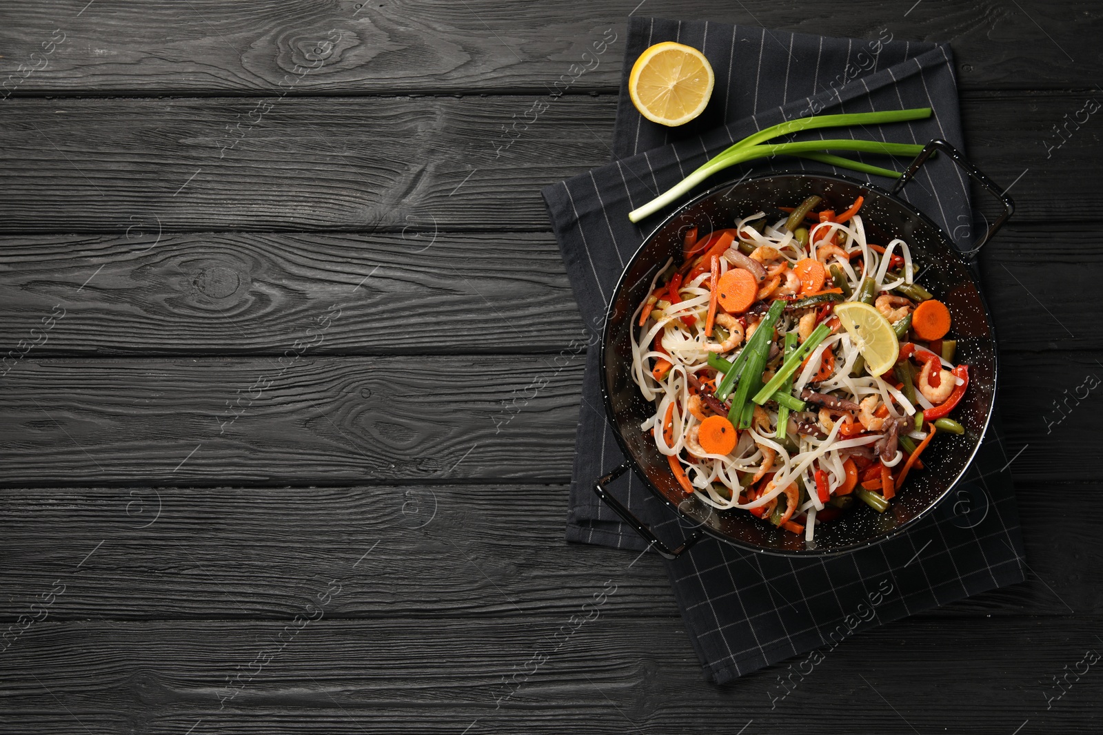 Photo of Shrimp stir fry with noodles and vegetables in wok on black wooden table, top view. Space for text