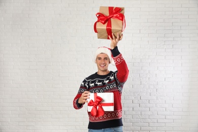 Photo of Happy man in Christmas sweater and Santa hat holding gift boxes near white brick wall