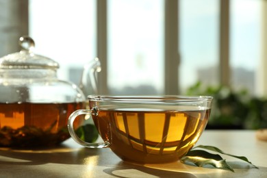 Photo of Fresh green tea in glass cup, leaves and teapot on table indoors