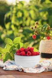 Photo of Tasty ripe raspberries in bowl, green leaves and straw hat on wooden table outdoors, space for text