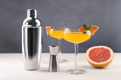 Photo of Metal shaker, delicious cocktail in glasses, jigger and grapefruit on light table