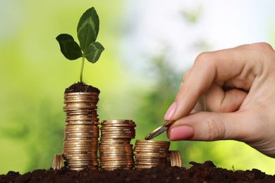 Photo of Woman putting coin onto stack with green sprout on soil against blurred background, closeup. Investment concept