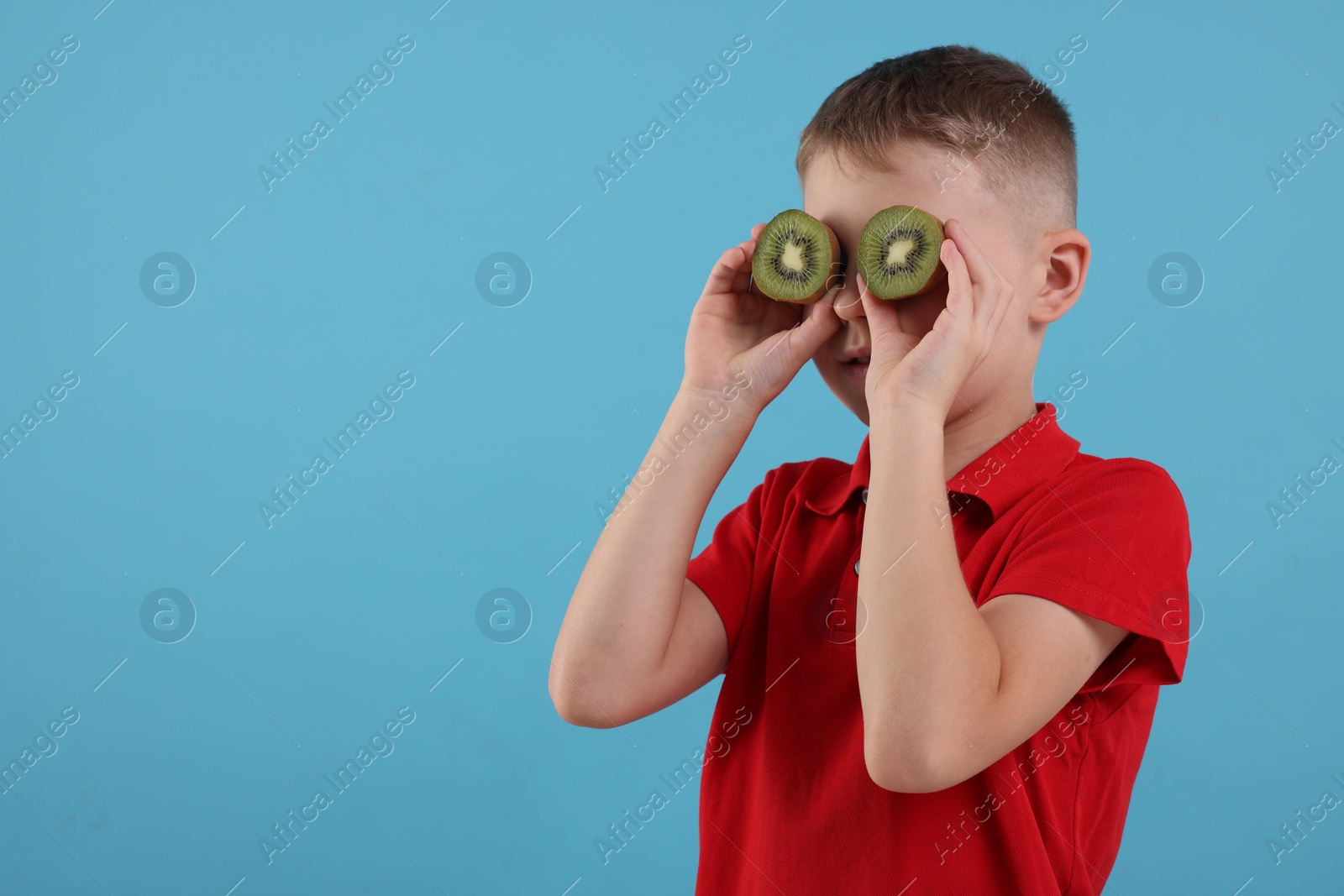 Photo of Boy covering eyes with halves of fresh kiwi on light blue background, space for text