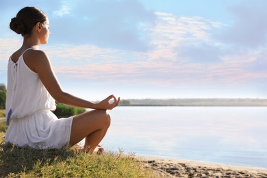 Young woman meditating near river at sunset, space for text. Nature healing power