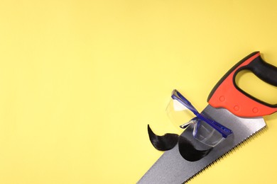 Photo of Man's face made of artificial mustache, safety glasses and hand saw on yellow background, top view. Space for text