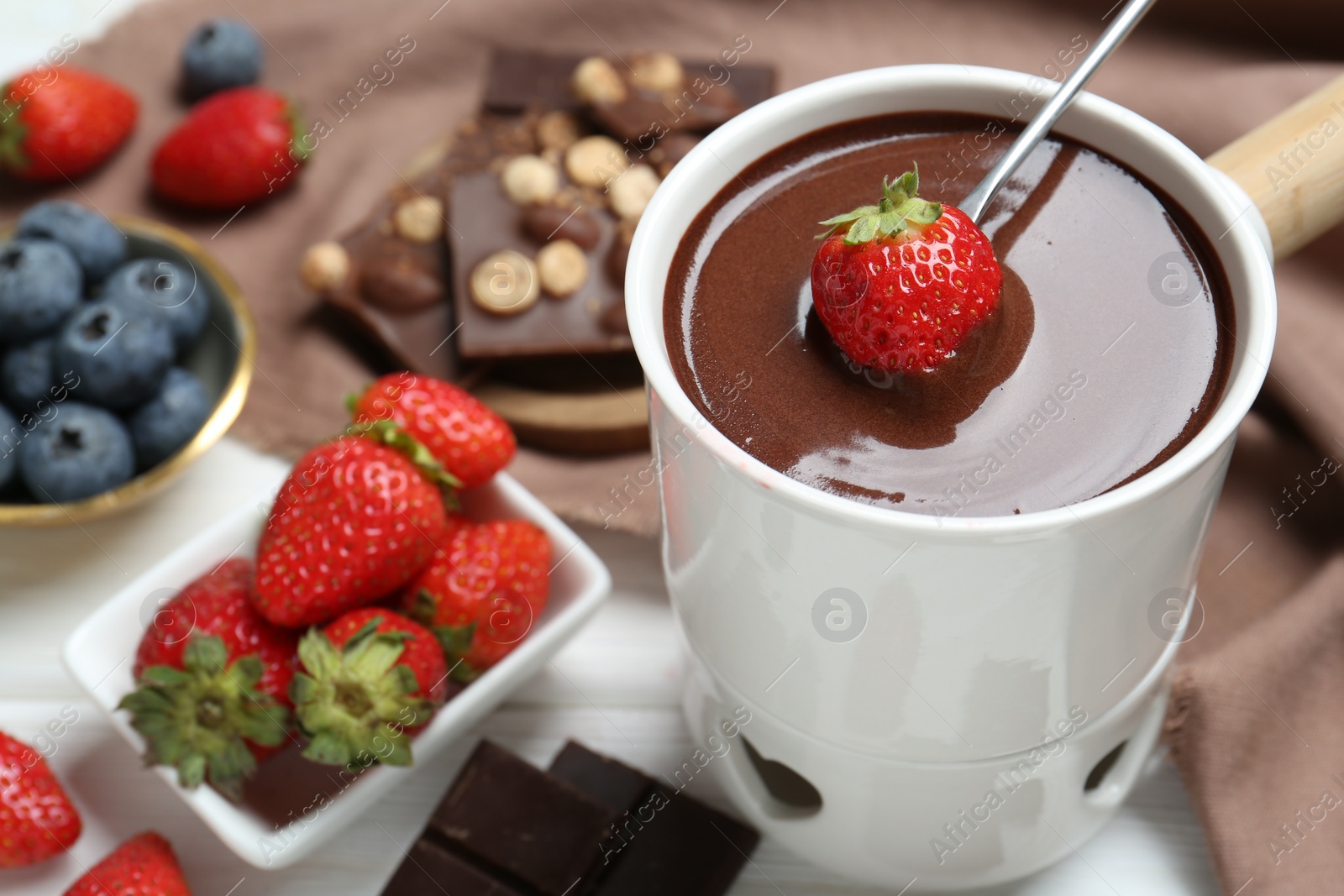 Photo of Dipping fresh strawberry in fondue pot with melted chocolate at white table, closeup