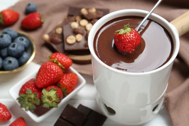 Photo of Dipping fresh strawberry in fondue pot with melted chocolate at white table, closeup