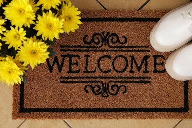 Photo of Door mat with word Welcome, stylish boots and beautiful flowers on floor, flat lay