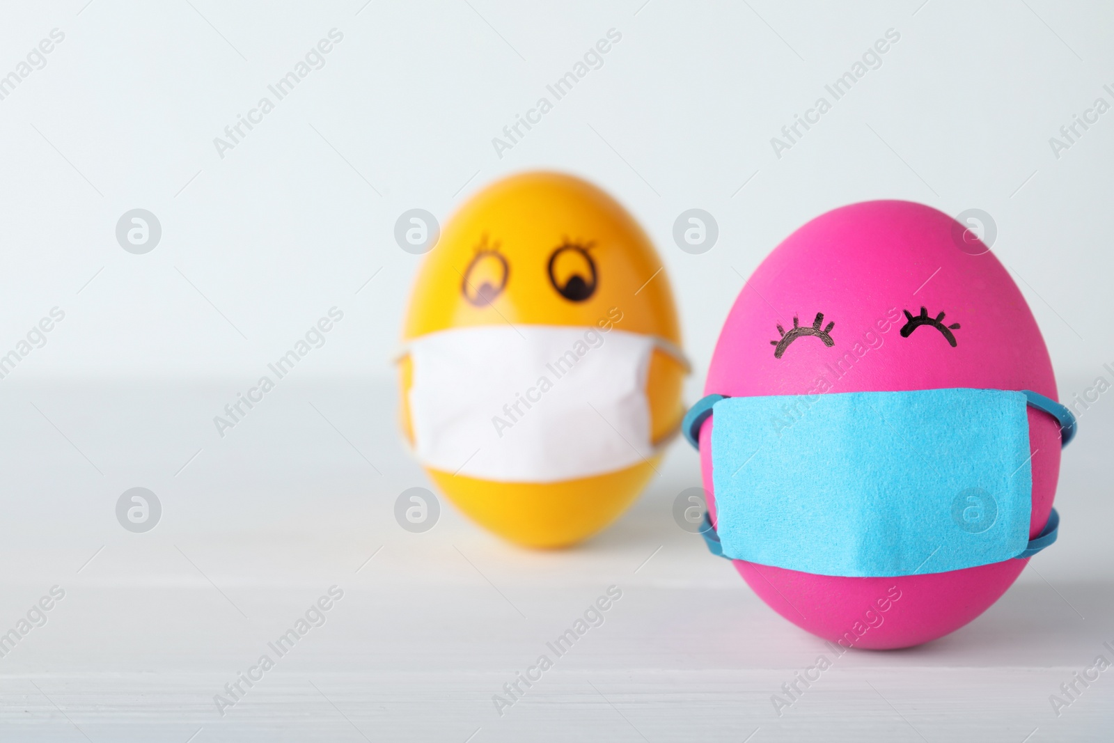 Photo of Bright eggs in protective masks on white wooden table, space for text. Easter holiday during coronavirus quarantine