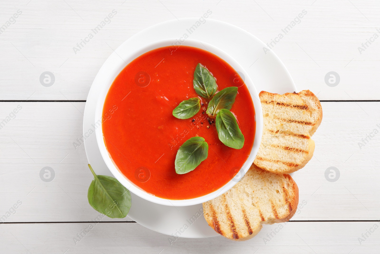 Photo of Delicious tomato cream soup in bowl with pieces of grilled bread on white wooden table, top view