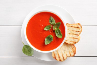 Delicious tomato cream soup in bowl with pieces of grilled bread on white wooden table, top view