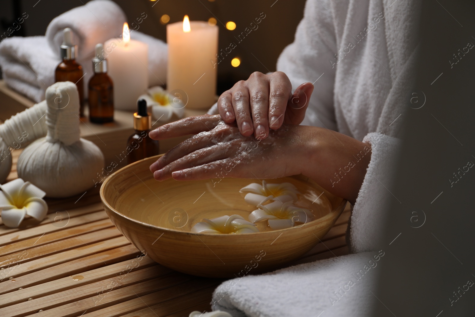 Photo of Woman applying scrub onto her hands in spa, closeup. Bowl of water and flowers on wooden table