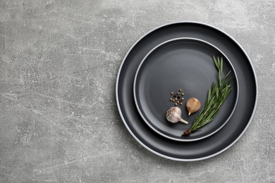 Photo of New dark plates with rosemary, garlic and peppercorns on light grey table, top view. Space for text