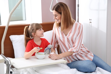 Photo of Little child with intravenous drip eating soup in hospital bed during mother's visit