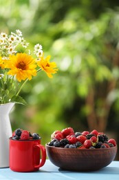 Photo of Different fresh ripe berries and beautiful flowers on light blue table outdoors