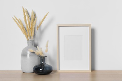 Photo of Empty photo frame and vases with dry decorative spikes on wooden table. Mockup for design