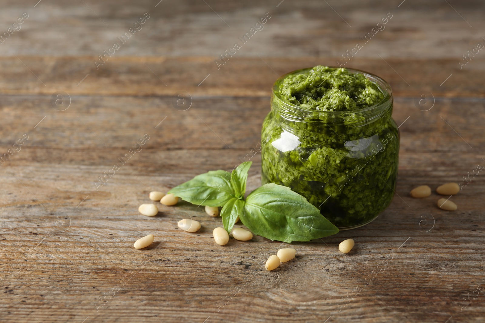 Photo of Jar of pesto sauce with basil leaves and pine nuts on wooden table. Space for text