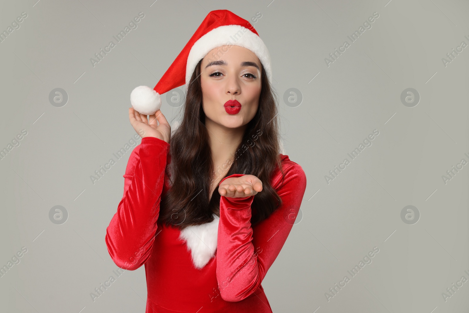 Photo of Christmas celebration. Beautiful young woman in red dress and Santa hat blowing kiss on grey background, space for text