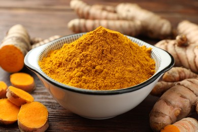 Bowl with aromatic turmeric powder and cut roots on wooden table, closeup