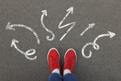 Choosing future profession. Girl standing in front of drawn signs on asphalt, top view. Arrows pointing in different directions symbolizing diversity of opportunities