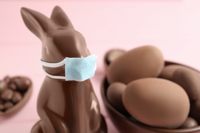 Chocolate bunny with protective mask and eggs on pink background, closeup. Easter holiday during COVID-19 quarantine