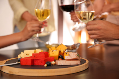 Different delicious cheeses on table and blurred friends toasting with wine on background