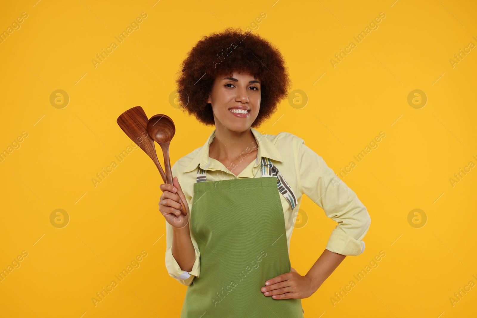 Photo of Happy young woman in apron holding spoon and spatula on orange background