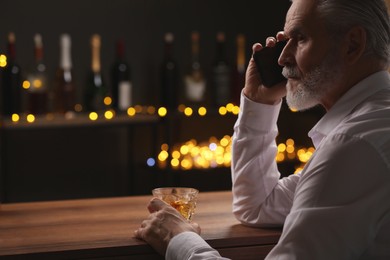 Photo of Senior man with glass of whiskey talking on phone at bar counter against blurred lights. Space for text