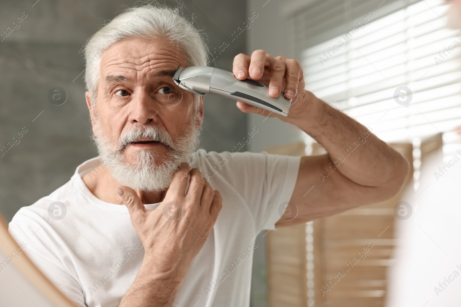 Photo of Senior man trimming eyebrows near mirror in bathroom