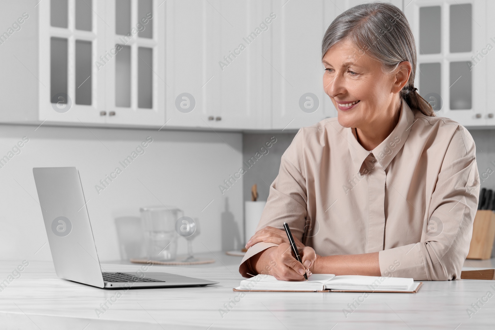 Photo of Beautiful senior woman writing something in notebook while using laptop at white marble table indoors