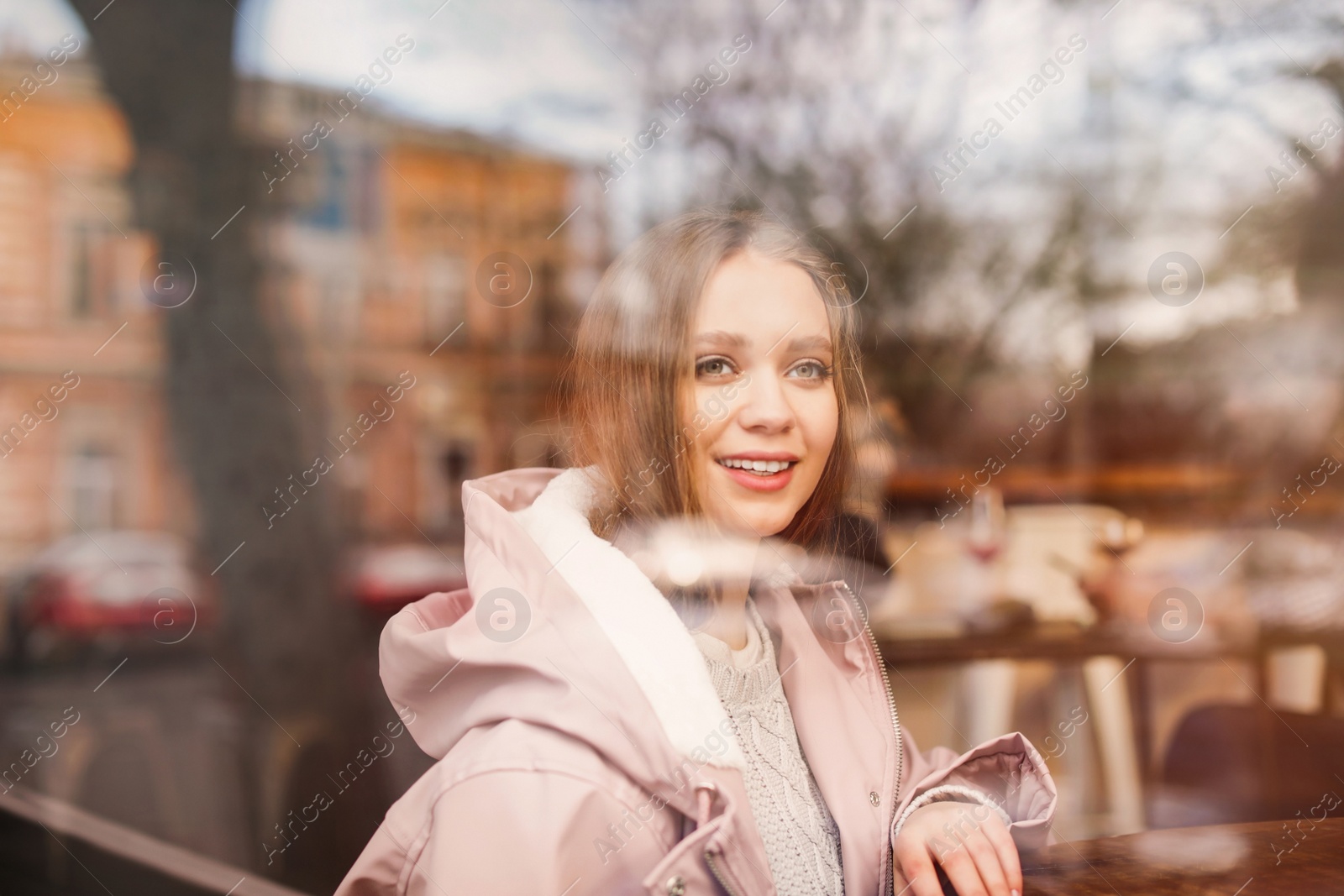 Photo of Beautiful young woman sitting at table in cafe, view from outdoors through window