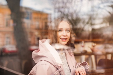 Beautiful young woman sitting at table in cafe, view from outdoors through window