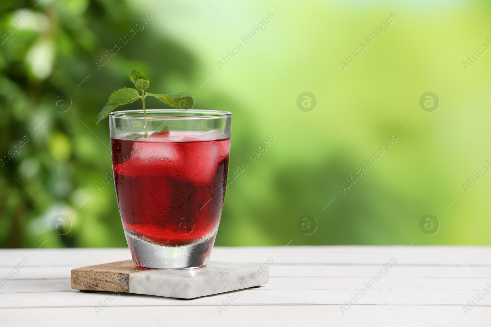 Photo of Refreshing hibiscus tea with ice cubes and mint in glass on white wooden table against blurred green background. Space for text