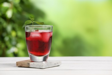 Photo of Refreshing hibiscus tea with ice cubes and mint in glass on white wooden table against blurred green background. Space for text