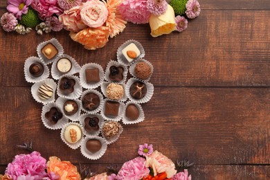 Photo of Heart made with delicious chocolate candies and beautiful flowers on wooden table, flat lay. Space for text