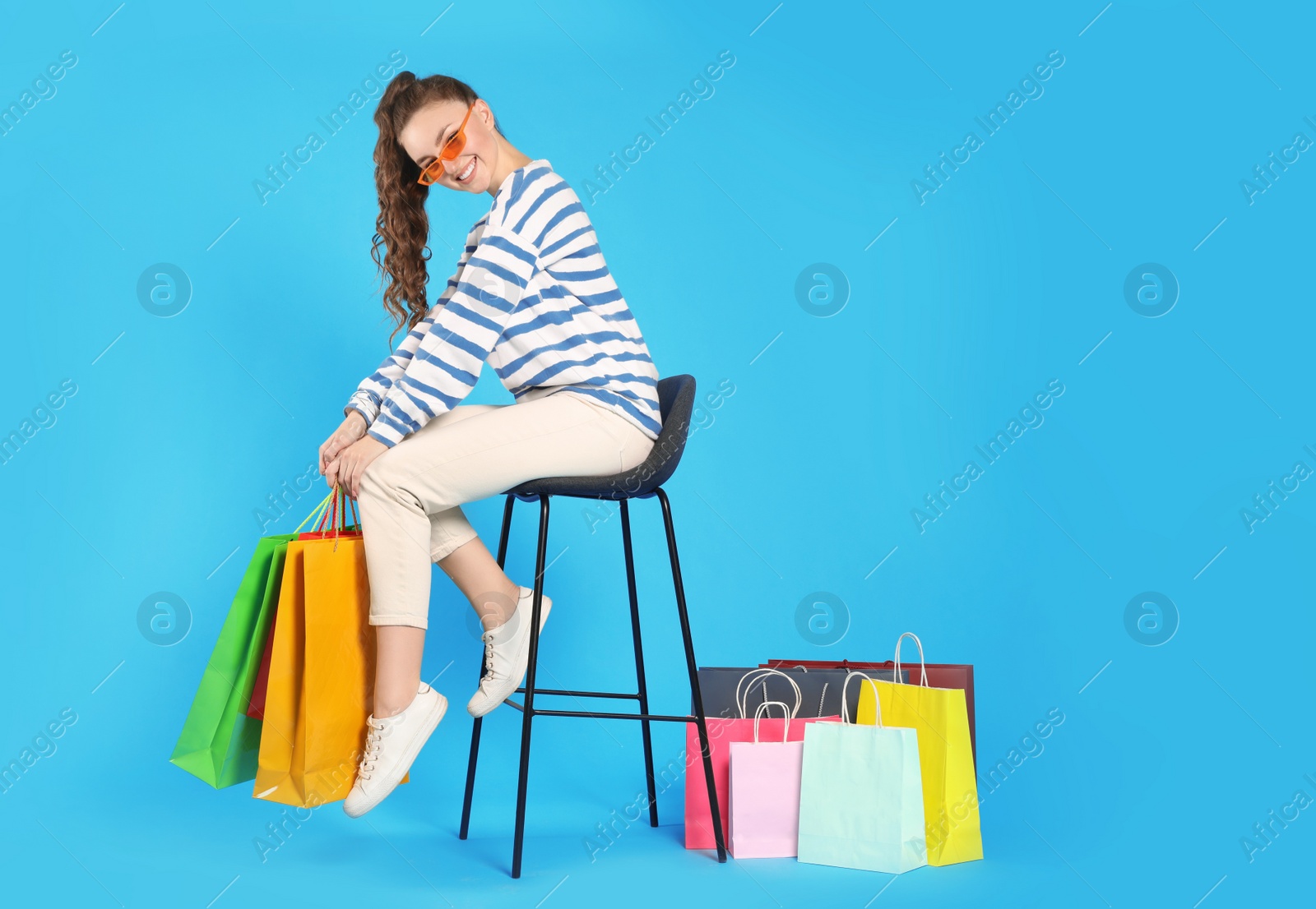 Photo of Happy woman in stylish sunglasses holding colorful shopping bags on stool against light blue background