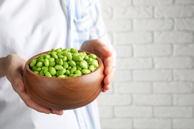 Photo of Woman holding bowl of edamame beans near white brick wall, closeup. Space for text