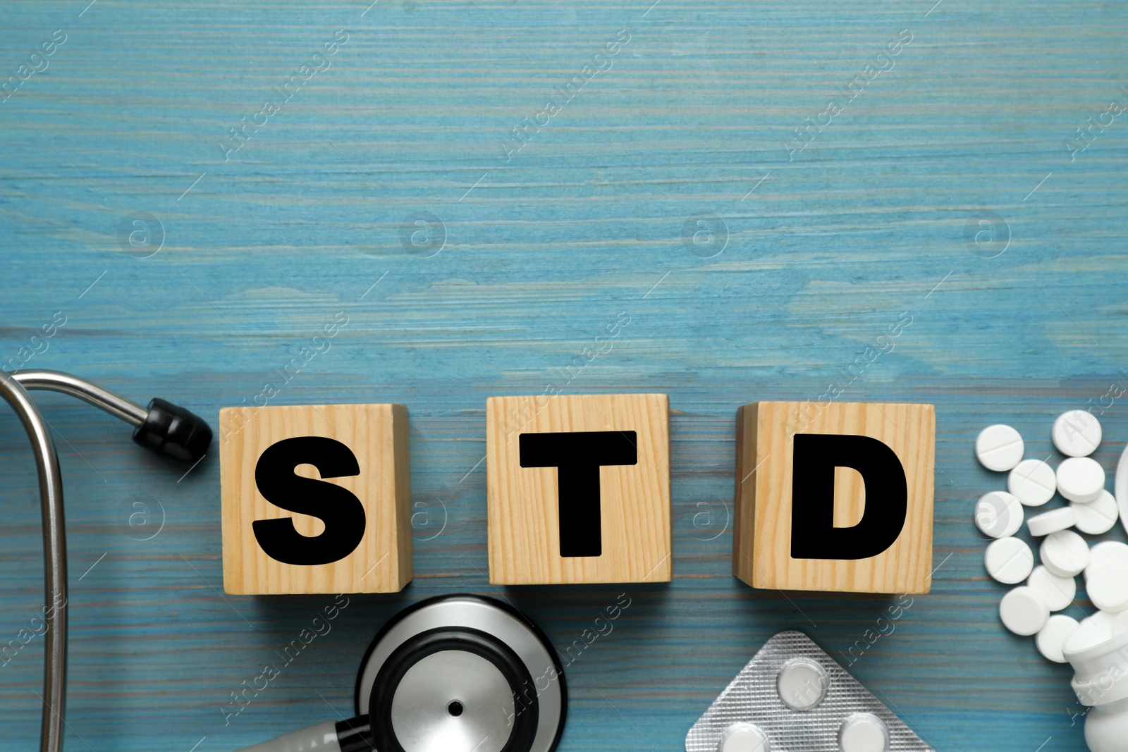 Photo of Abbreviation STD made with cubes, pills and stethoscope on light blue wooden table, flat lay. Space for text