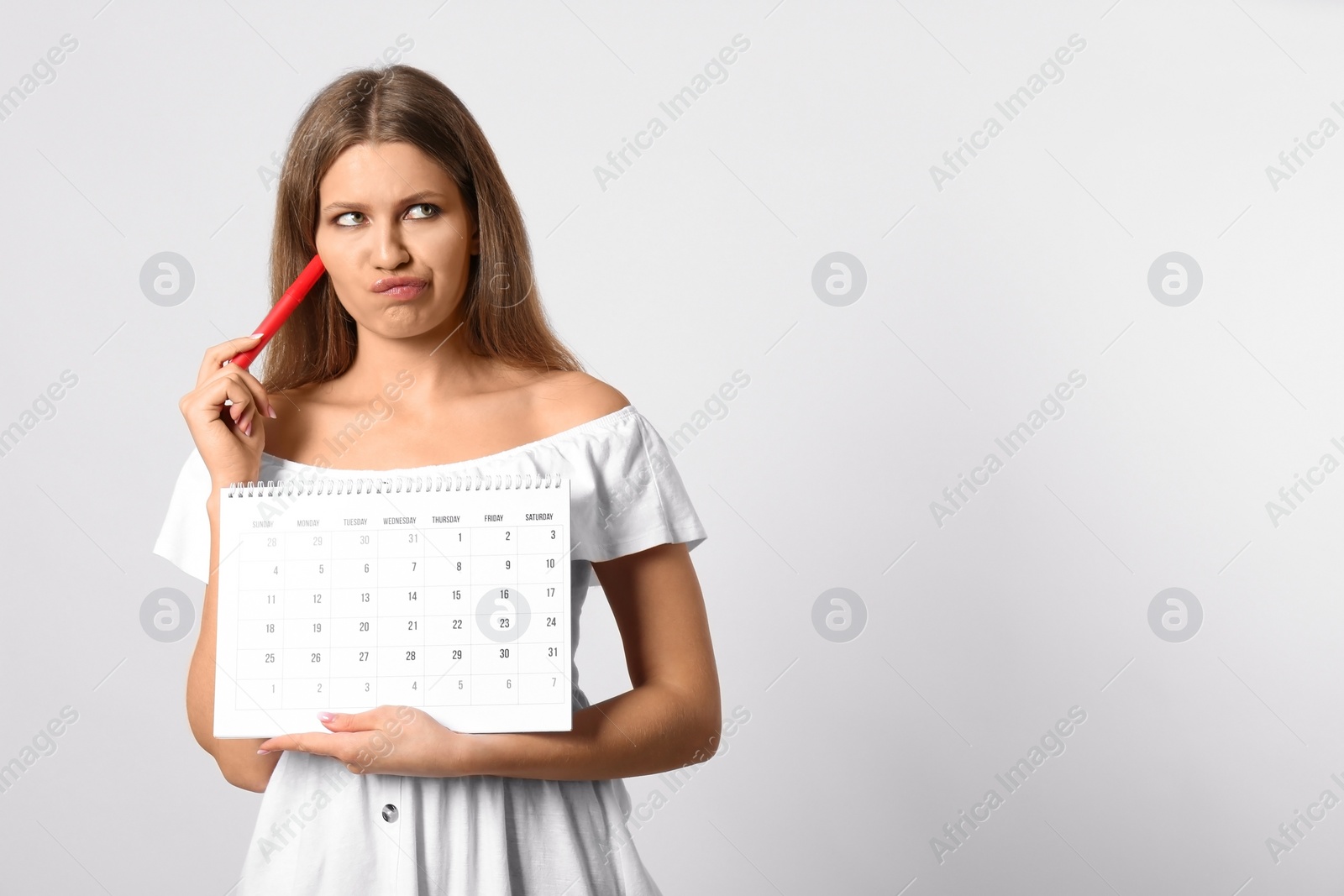 Photo of Pensive young woman holding calendar with marked menstrual cycle days on light background. Space for text
